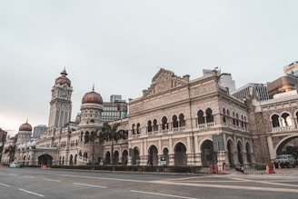 A historic and ornate building with a large clock tower and intricate architectural details dominates the scene, surrounded by palm trees and a wide street. The structure features arched windows and reddish domes, combining colonial and Moorish architectural styles.