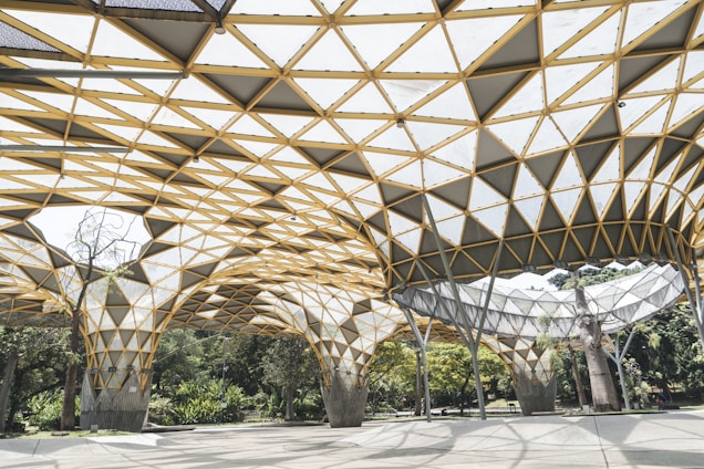 A geometric roof structure with a pattern of triangular shapes and a mix of glass and metal elements covers an outdoor area. The design features intersecting lines creating a honeycomb-like appearance. Beneath the structure, trees and greenery are visible, suggesting the setting is a blend of architecture and natural landscape.