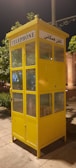 A bright yellow telephone booth with glass windows standing on a paved area. The booth has signs in English and another language, indicating its function as a telephone booth. There are green trees and some lit structures in the background, suggesting it might be in a park or an urban area.