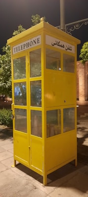 A bright yellow telephone booth with glass windows standing on a paved area. The booth has signs in English and another language, indicating its function as a telephone booth. There are green trees and some lit structures in the background, suggesting it might be in a park or an urban area.