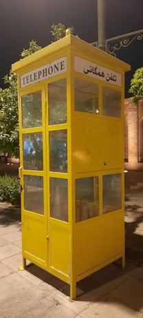 A bright yellow telephone booth with glass windows standing on a paved area. The booth has signs in English and another language, indicating its function as a telephone booth. There are green trees and some lit structures in the background, suggesting it might be in a park or an urban area.