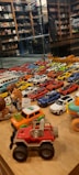 A collection of various toy cars and figurines displayed on a wooden surface. The cars are arranged in rows, showcasing a variety of colors including red, blue, white, and yellow. In the background, shelves filled with books and more toys can be seen, reflected in a glass partition.