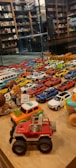 A collection of various toy cars and figurines displayed on a wooden surface. The cars are arranged in rows, showcasing a variety of colors including red, blue, white, and yellow. In the background, shelves filled with books and more toys can be seen, reflected in a glass partition.