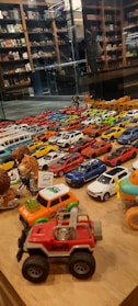 A collection of various toy cars and figurines displayed on a wooden surface. The cars are arranged in rows, showcasing a variety of colors including red, blue, white, and yellow. In the background, shelves filled with books and more toys can be seen, reflected in a glass partition.