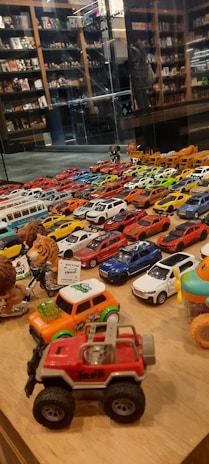 A collection of various toy cars and figurines displayed on a wooden surface. The cars are arranged in rows, showcasing a variety of colors including red, blue, white, and yellow. In the background, shelves filled with books and more toys can be seen, reflected in a glass partition.