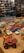 A collection of various toy cars and figurines displayed on a wooden surface. The cars are arranged in rows, showcasing a variety of colors including red, blue, white, and yellow. In the background, shelves filled with books and more toys can be seen, reflected in a glass partition.