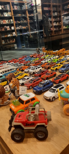 A collection of various toy cars and figurines displayed on a wooden surface. The cars are arranged in rows, showcasing a variety of colors including red, blue, white, and yellow. In the background, shelves filled with books and more toys can be seen, reflected in a glass partition.