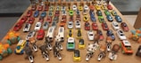 A collection of various toy cars and animal-themed figurines arranged neatly on a wooden table. The toy cars are organized in rows, showcasing a wide range of colors including red, blue, yellow, orange, white, and green. At the forefront, there are figurines resembling animals such as elephants, tigers, pandas, and lions, each riding a small motorcycle. The background features toy trucks. The arrangement suggests a carefully curated display, possibly for decorative or collectible purposes.