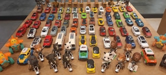 A collection of various toy cars and animal-themed figurines arranged neatly on a wooden table. The toy cars are organized in rows, showcasing a wide range of colors including red, blue, yellow, orange, white, and green. At the forefront, there are figurines resembling animals such as elephants, tigers, pandas, and lions, each riding a small motorcycle. The background features toy trucks. The arrangement suggests a carefully curated display, possibly for decorative or collectible purposes.