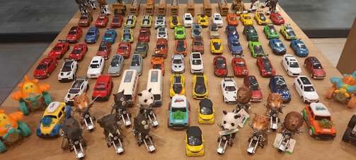 A collection of various toy cars and animal-themed figurines arranged neatly on a wooden table. The toy cars are organized in rows, showcasing a wide range of colors including red, blue, yellow, orange, white, and green. At the forefront, there are figurines resembling animals such as elephants, tigers, pandas, and lions, each riding a small motorcycle. The background features toy trucks. The arrangement suggests a carefully curated display, possibly for decorative or collectible purposes.
