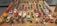 A collection of various toy cars and animal-themed figurines arranged neatly on a wooden table. The toy cars are organized in rows, showcasing a wide range of colors including red, blue, yellow, orange, white, and green. At the forefront, there are figurines resembling animals such as elephants, tigers, pandas, and lions, each riding a small motorcycle. The background features toy trucks. The arrangement suggests a carefully curated display, possibly for decorative or collectible purposes.
