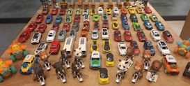 A collection of various toy cars and animal-themed figurines arranged neatly on a wooden table. The toy cars are organized in rows, showcasing a wide range of colors including red, blue, yellow, orange, white, and green. At the forefront, there are figurines resembling animals such as elephants, tigers, pandas, and lions, each riding a small motorcycle. The background features toy trucks. The arrangement suggests a carefully curated display, possibly for decorative or collectible purposes.
