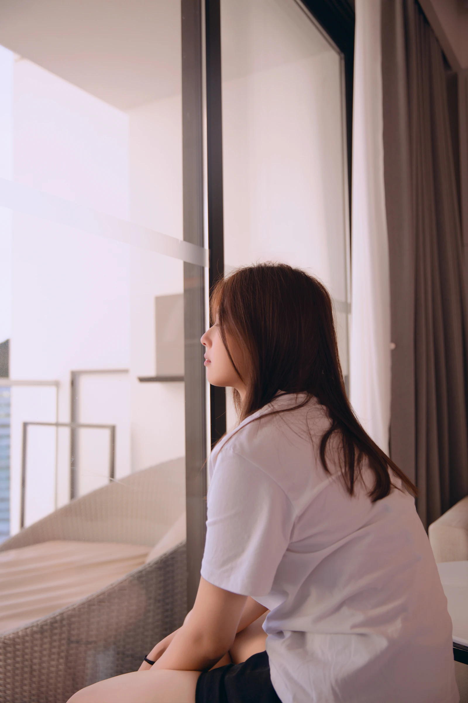A woman sitting quietly on the edge of a bed by a window, attention turned inward