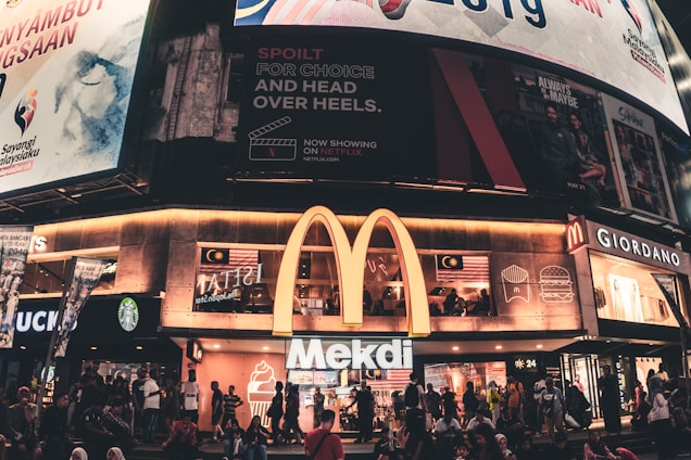 A busy urban scene featuring a McDonald's restaurant with a large illuminated 'M' logo. Above the restaurant are several large billboards displaying various advertisements, including a Netflix promo and Malaysian national imagery. People are gathered outside on the sidewalk, some sitting and others walking by. Nearby shops include a Starbucks and a Giordano.