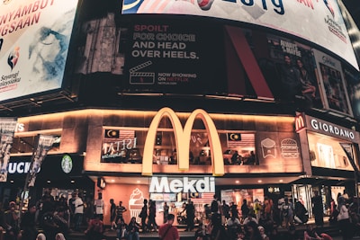 A busy urban scene featuring a McDonald's restaurant with a large illuminated 'M' logo. Above the restaurant are several large billboards displaying various advertisements, including a Netflix promo and Malaysian national imagery. People are gathered outside on the sidewalk, some sitting and others walking by. Nearby shops include a Starbucks and a Giordano.
