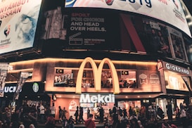A busy urban scene featuring a McDonald's restaurant with a large illuminated 'M' logo. Above the restaurant are several large billboards displaying various advertisements, including a Netflix promo and Malaysian national imagery. People are gathered outside on the sidewalk, some sitting and others walking by. Nearby shops include a Starbucks and a Giordano.