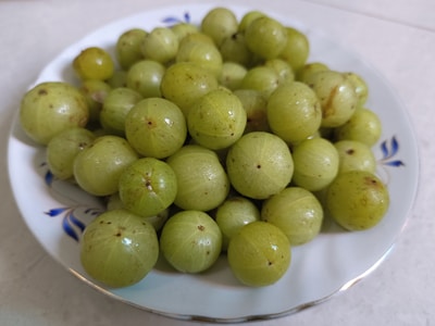 A white plate filled with a pile of green Indian gooseberries, also known as amla. The berries are round with a slightly shiny surface, some showing small brown spots. The plate is placed on a light-colored surface.
