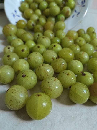 A collection of fresh green Indian gooseberries scattered across a light-colored surface, with some falling out of a white bowl adorned with blue floral designs.