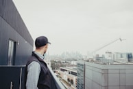 A person wearing a cap stands on a rooftop, gazing at a distant city skyline. A large construction crane is visible to the right, and several modern buildings populate the view.