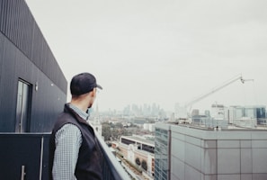 A person wearing a cap stands on a rooftop, gazing at a distant city skyline. A large construction crane is visible to the right, and several modern buildings populate the view.