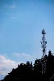 A communication tower stands tall against a clear blue sky, partially bordered by the silhouettes of lush trees.