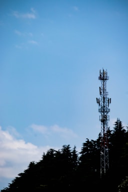 A communication tower stands tall against a clear blue sky, partially bordered by the silhouettes of lush trees.