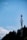 A communication tower stands tall against a clear blue sky, partially bordered by the silhouettes of lush trees.