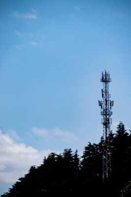 A communication tower stands tall against a clear blue sky, partially bordered by the silhouettes of lush trees.