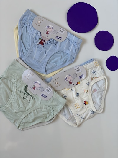 Three pairs of children's underwear are laid out on a flat surface. The underwear has playful designs featuring cartoon characters and patterns. There are three large purple circles placed next to the underwear.