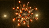A symmetrical arrangement of small lit candles is placed on a dark, glossy surface. The candles form a circular pattern around intricate white line designs that resemble traditional rangoli art.