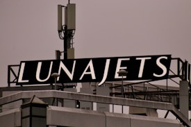 A rooftop structure featuring a large black sign with the word 'LUNAJETS' in bold white letters. The sign is mounted above an industrial-looking building with a complex array of antennas and metal railings.