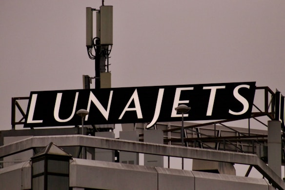 A rooftop structure featuring a large black sign with the word 'LUNAJETS' in bold white letters. The sign is mounted above an industrial-looking building with a complex array of antennas and metal railings.