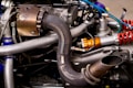 An intricate assembly of a car engine with visible exhaust pipes, a turbocharger, and various connecting rods and wires. The components are metallic with a variety of finishes, including polished silver and copper-colored parts alongside colorful tubing in blue and orange.