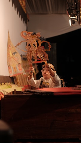A person is performing a traditional puppet show using intricately designed shadow puppets. The puppeteer is wearing traditional attire and holds the puppets aloft in a dramatic pose. The setting is dimly lit, emphasizing the warm tones and intricate details of the puppets and backdrop.