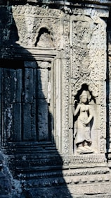 Stone carvings on an ancient temple wall are intricately detailed, featuring a relief sculpture of a standing woman. The detailed patterns and decorative elements surround the figure, with natural shadows cast across the stone surface. The craftsmanship reflects historical artistic styles.