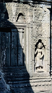 Stone carvings on an ancient temple wall are intricately detailed, featuring a relief sculpture of a standing woman. The detailed patterns and decorative elements surround the figure, with natural shadows cast across the stone surface. The craftsmanship reflects historical artistic styles.