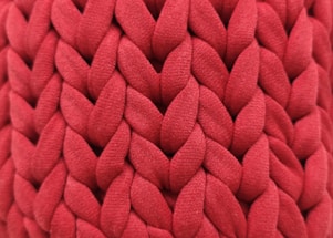 A close-up view of an intricate red knitted fabric with a thick, chunky texture. The woven pattern displays a series of large, interlocking loops creating a visually rich surface.