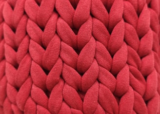 A close-up view of an intricate red knitted fabric with a thick, chunky texture. The woven pattern displays a series of large, interlocking loops creating a visually rich surface.