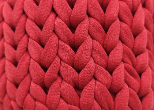 A close-up view of an intricate red knitted fabric with a thick, chunky texture. The woven pattern displays a series of large, interlocking loops creating a visually rich surface.