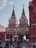 A group of people walking around a historic gate with two tall, ornate spires. The gate features intricate architectural details in red and white, along with a central archway topped by a decorative blue and gold element. The scene appears to be bustling with activity.