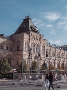 A historic building with intricate architectural details is in the background, featuring ornate facades and a steep roof. In the foreground, people are assembling scaffolding, likely part of a construction or maintenance project. Lush green trees line the scene, and passersby walk along the cobblestone path, suggesting an active urban environment.
