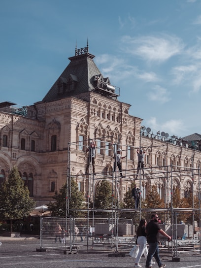 A historic building with intricate architectural details is in the background, featuring ornate facades and a steep roof. In the foreground, people are assembling scaffolding, likely part of a construction or maintenance project. Lush green trees line the scene, and passersby walk along the cobblestone path, suggesting an active urban environment.