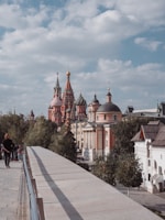 A scenic view featuring colorful, ornate domes of a famous building in Moscow, surrounded by trees and other classical architecture. A few people are walking along a pathway lined with trees and buildings on a clear day with scattered clouds.