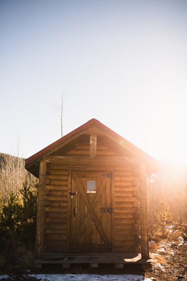 A small wooden cabin is situated in a natural setting with a clear blue sky and the sun shining brightly, casting a warm glow. The cabin has a slanted roof with a red trim and a door made of wood with a small window on it. Sparse vegetation surrounds the cabin, with a few bushes and patches of snow on the ground.