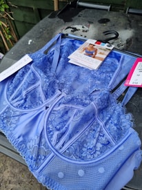 A vibrant blue lace lingerie set displayed on a hanger. The intricate floral lace design covers the fabric, and there is a product tag featuring an image and details about the lingerie. The background shows an outdoor setting with some greenery.