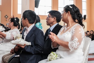 A group of people dressed formally, possibly attending a ceremony. The woman in the foreground is wearing a white lace dress and glasses, while the man beside her is wearing a dark suit. A young boy is also visible, holding a white cushion with small decorative items.
