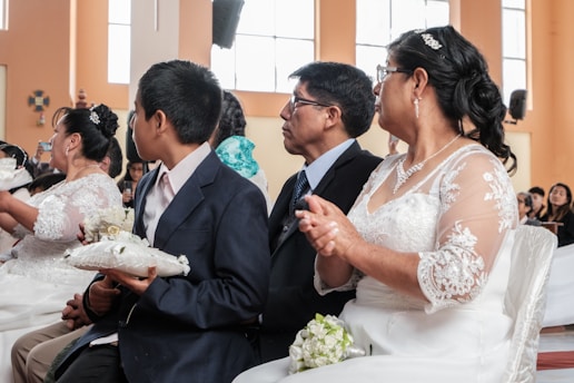 A group of people dressed formally, possibly attending a ceremony. The woman in the foreground is wearing a white lace dress and glasses, while the man beside her is wearing a dark suit. A young boy is also visible, holding a white cushion with small decorative items.