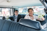 A couple is sitting in the backseat of a car, dressed in wedding attire. The woman is holding a bouquet of white flowers and wearing a lace dress with a necklace, while the man is in a suit with a tie, both smiling. In the background, there are people outside the car and other vehicles visible through the windows.