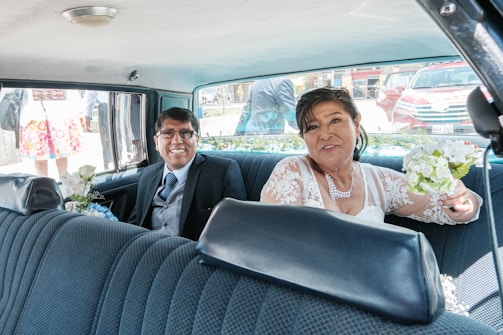 A couple is sitting in the backseat of a car, dressed in wedding attire. The woman is holding a bouquet of white flowers and wearing a lace dress with a necklace, while the man is in a suit with a tie, both smiling. In the background, there are people outside the car and other vehicles visible through the windows.