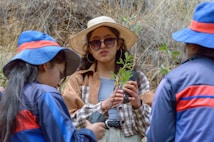Three people are engaged in planting activities outdoors, with one holding a small plant. The individual in the middle wears a large sun hat and sunglasses, while the others wear blue hats and jackets with red accents. The background features dry grasses and plants.